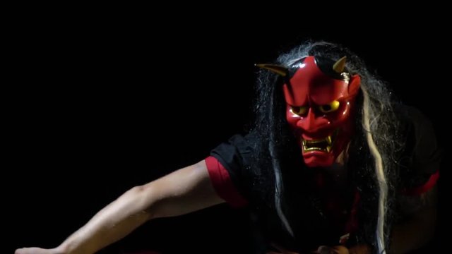 A freak in a black kimono hides a face behind a red demon mask with long hair and flaps a red fan