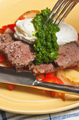Steak, peppers and eggs with arugula pesto on a round, yellow plate with a fork about to pierce the egg and a knife on a blue and white table cloth paleo diet
