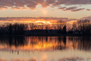 Sunset on a lake, with symmetric reflections of trees silhouettes and clouds on water