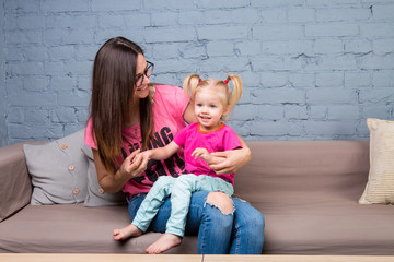 Mom and baby are playing and having fun in the room on the couch. They are dressed in bright clothes. Attractive, stylish and young family