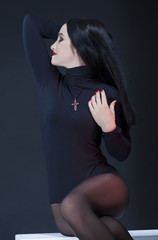 Studio Portrait of Passionate Sexy Caucasian Brunette Woman in Black Body Suit Posing on White Box Against Black Background.