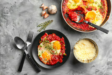 Plate and frying pan with eggs in purgatory on grey background