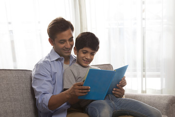 Smiling father and son reading book sitting on sofa