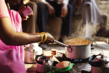 Cropped image of female vendor preparing chai on stove with customers in background 