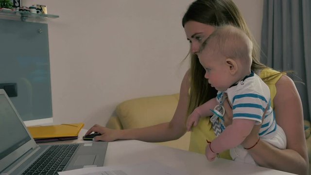 Tired mother works with laptop with baby in arms