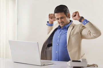 Tired Indian businessman stretching with laptop at office desk