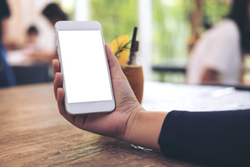 Mockup image of a woman's hand holding white mobile phone with blank screen with a glass of iced coffee on wooden table in cafe