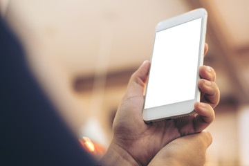 Mockup image of hands holding and raising white mobile phone with blank screen in vintage cafe
