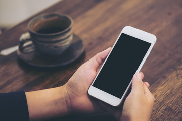 Mockup image of hand holding white mobile phone with blank black screen and coffee cup on vintage wooden table