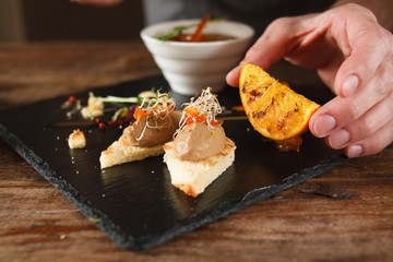 Culinary art of serving appetizers in restaurant. Unrecognizable man lays grilled orange and herbs near small canapes with paste on black tray