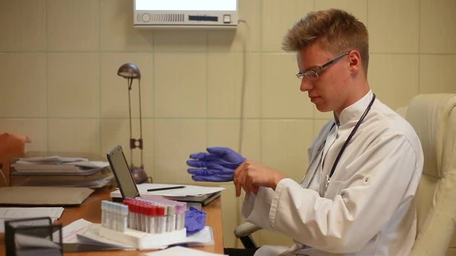 Doctor sitting in the office by the desk and put medical gloves on
