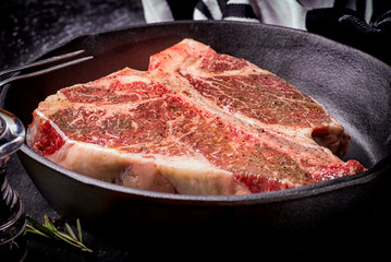 Fresh raw meat t-bone steak in a skillet, marinated with herbs and spices, shot with a dark background.