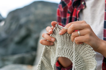 Close-up Young woman checkered red shirt and white t-shirt knits with knitting needles gray sweater from natural woolen threads, in the background stones