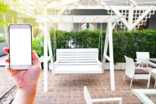 Hand holding smartphone on the white chair and table in the green garden with white swing background - Meeting point of concept