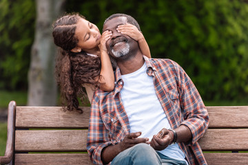 smiling african american girl closing eyes of her grandfather sitting on bench in park