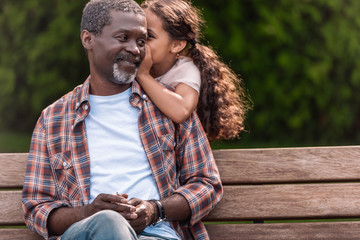 little adorable african american girl whispering to her grandfather while sitting on bench in park