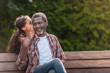 little adorable african american girl whispering to her grandfather while sitting on bench in park