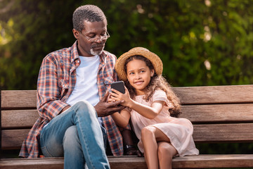 smiling african american girl and her grandfather using smartphone sitting on bench together