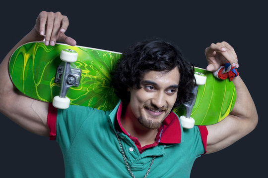 Close-up of young smiling man holding skateboard behind head against black background 