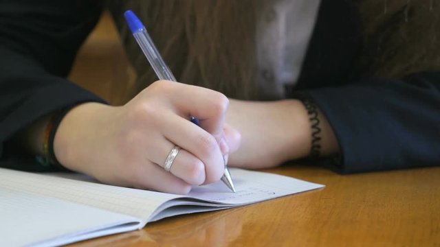 Schoolgirl dressed in a black suit sits at a school desk writing text in exercise book using ballpoint pen. Close-up