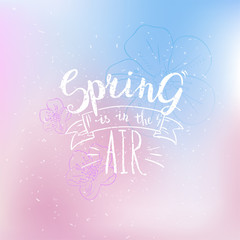 Spring is in the air card. Hand drawn calligraphy phrase on abstract blurred background with  flowers. Pastel stylish spring backdrop