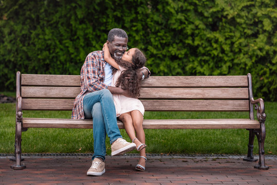 adorable african american girl hugging and kissing her grandfather while sitting on bench in park - Powered by Adobe