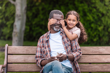 smiling african american girl closing eyes of her grandfather sitting on bench in park