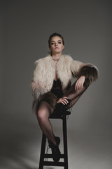 seductive fashionable young girl in black sexy clothes and fur coat sitting on stool on grey