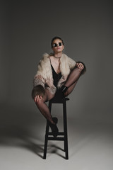 Fototapeta na wymiar seductive fashionable young girl in sunglasses, black sexy clothes and fur coat sitting on stool on grey