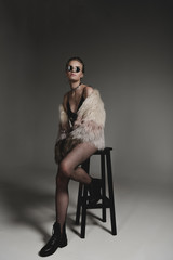 Fototapeta na wymiar attractive fashionable young girl in sunglasses, black sexy clothes and fur coat sitting on stool on grey