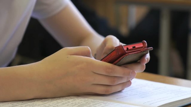 Schoolgirl sitting at the school desk at school writes the text using a red mobile phone indoors