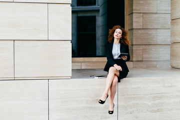 Lovely curly woman with confident look, sitting alone, demonstrating her slender long legs, dressed in black jacket with skirt, holding tablet, looking schedules or diagrams, making business report