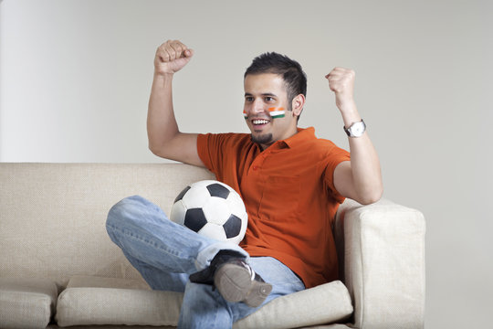 Happy young man with soccer ball cheering while sitting on sofa 