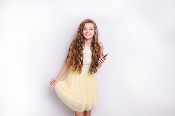 Cute blond girl listening to music on earphones on smartphone is having fun. She smiles widely and plays with her yellow dress. She had long curly blond hair.