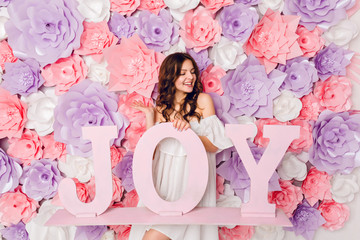 Vertical portrait of cute brunette girl. She stands and holds wood word JOY smiling widely. She has curly hair and wears white dress off the shoulder. She has pink background covered in flowers.
