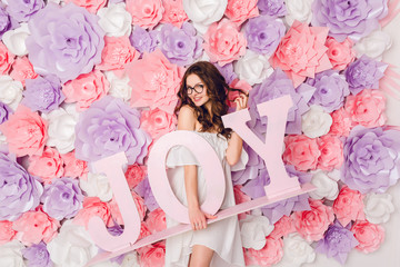 Vertical portrait of cute brunette girl. She stands and holds wood word JOY smiling widely. She has curly hair and wears white dress off the shoulder and black glasses. Background in pink flowers.