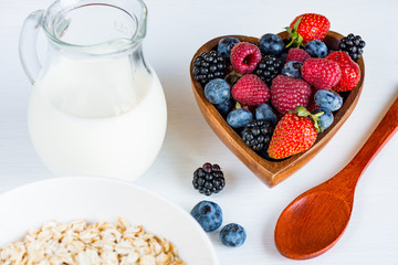 Berries of strawberries, raspberries, blackberries, blueberries in wooden bowl, spoon, jug of milk and muesli on a white wooden table