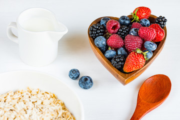 Berries of strawberries, raspberries, blackberries, blueberries in wooden bowl, spoon, jug of milk and muesli on a white wooden table