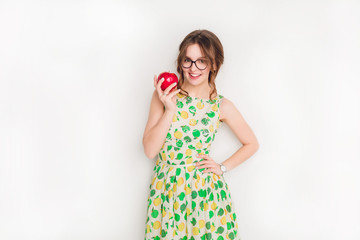 Studio shot of a smiling brunette girl smiling widely. Girl wears black glasses and yellow and green dress. She holds a red apple in her right hand.