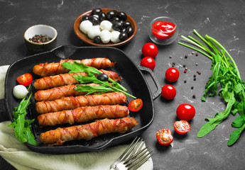 Preparation of roast grill sausages wrapped spirally in bacon on a cast-iron frying pan. Ingredients for sausages in bacon cherry tomatoes, rucola salad, pepper. Black concrete background. 