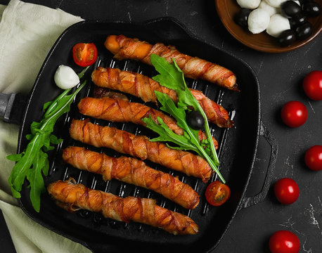 Preparation of roast grill sausages wrapped spirally in bacon on a cast-iron frying pan. Ingredients for sausages in bacon cherry tomatoes, rucola salad, pepper. Black concrete background. 