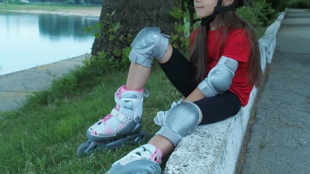 A girl on roller skates sits by the river. A child on rollers and a helmet resting on the river bank.