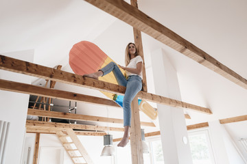 Carefree young woman sitting on ceiling joist, holding colorful airbed