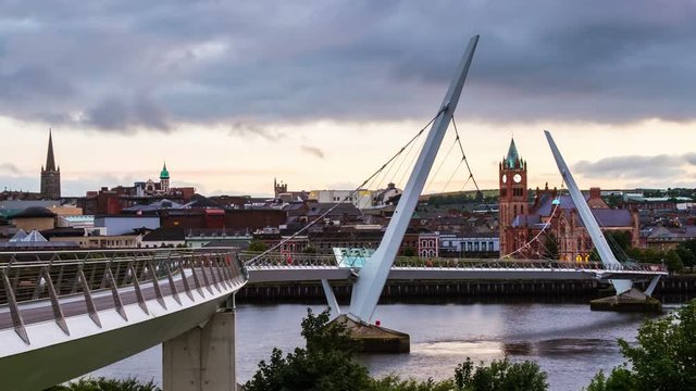 Derry, Ireland. Illuminated Peace bridge in Derry Londonderry in Northern Ireland with city center at the background. Time-lapse from day to night with cloudy sky, reflection in the river.