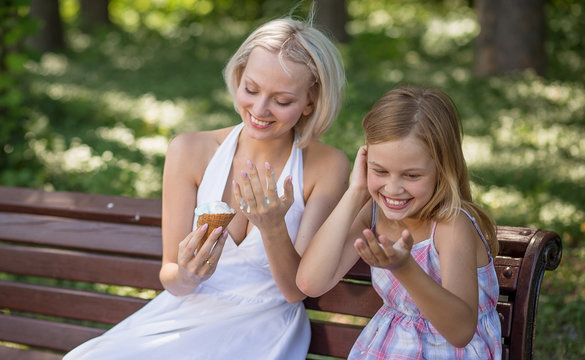 Young mother holding melting ice cream. laughing. Mom and daughter sitting on bench in the park, having ice cream.