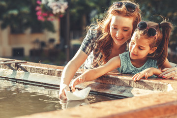 On a warm summer day, a mother, and her daughter joyfully set sail paper boats in a vibrant...