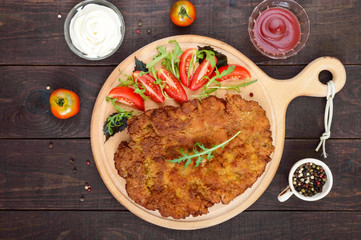 A large Viennese schnitzel  and tomato salad on a cutting board on a dark wooden background. Top view.