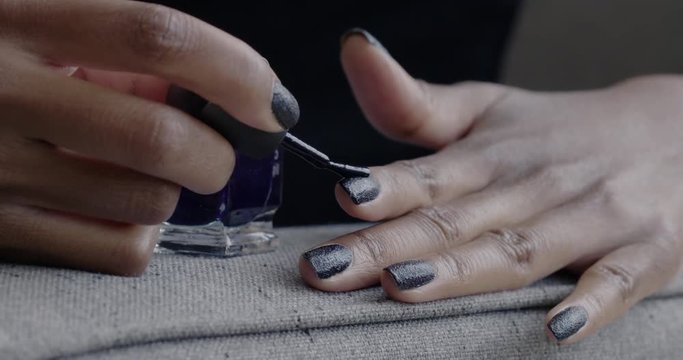 African American woman applies dark, glittering polish to a finger nail as she rests her hand on the arm of a sofa in natural light. Slow motion 4K, recorded at 60fps.