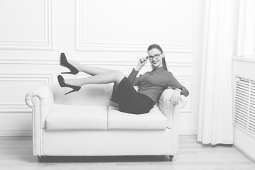 Attractive brunette businesswoman in eyeglasses lying on white sofa as symbol of sex, seduction, business, work, success, style, leadership, management, entrepreneur, Monochrome image