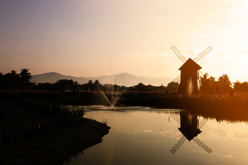 The silhouette of a windmill  on pond in the center of a flower garden with the sunshine in the evening.
